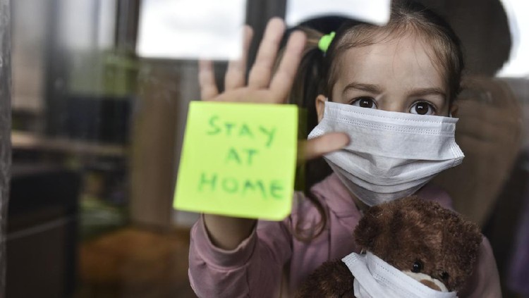 1218365817 Concept for social distancing during coronavirus pandemic. A young girl and his teddy bear are wearing protective facemarks while seeking protection from COVID-19. STAY HOME