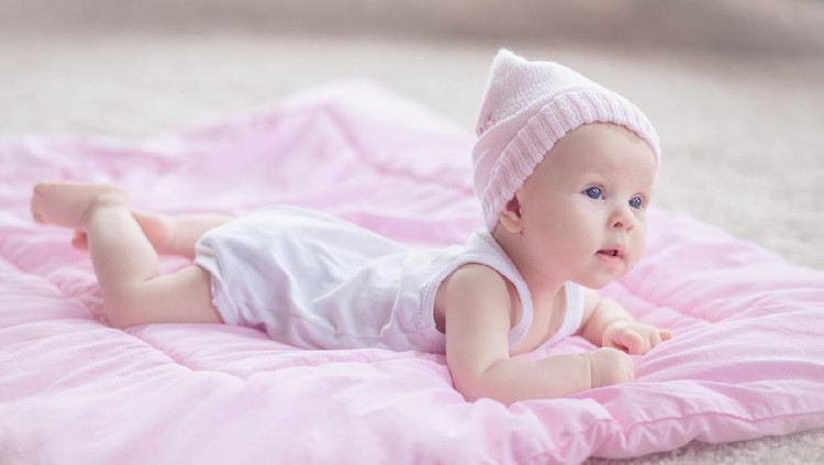 Cute playfull baby girl, wearing pink winter hat, lying at her tommy on a pink comforter on the floor. She's curious about her room interior.