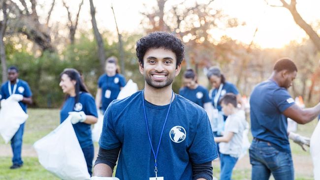 A young Indian man smiles at the camera while volunteering with his neighbors to pick up garbage in their neighborhood park. He is holding a garbage bag.