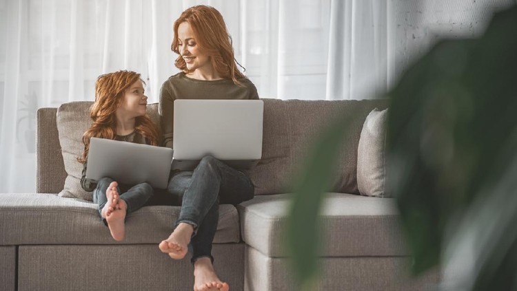 Full length portrait of delighted mom and kid sitting on sofa at home. They are holding computers while looking at each other with smile and love