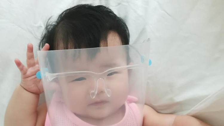 The baby wears a face shield lying on white bedsheet in pink vest.