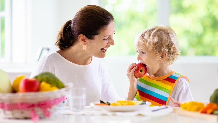 Mother feeding child vegetables. Mom feeds kid in white kitchen with window. Baby boy sitting in high chair eating healthy lunch of steamed carrot and broccoli. Nutrition, vegetarian diet for toddler