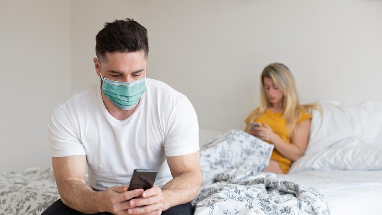 Couple under Covid-19 quarantine using their phones at home. Man wearing face mask texting on smartphone.