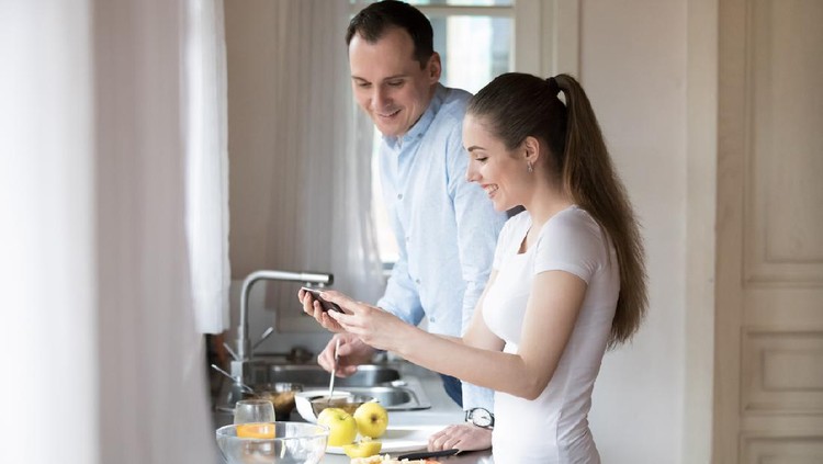 Happy young couple watching video on smartphone while cooking in kitchen at home. Attractive millennial wife laughing showing husband funny clip. Gadget addiction concept. People interacting at home