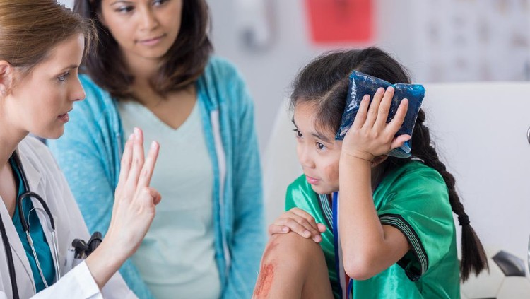 Young female soccer athlete is dazed while ER doctor asks her questions. The girl has an icepack on her head. The girl's mom is in the background.