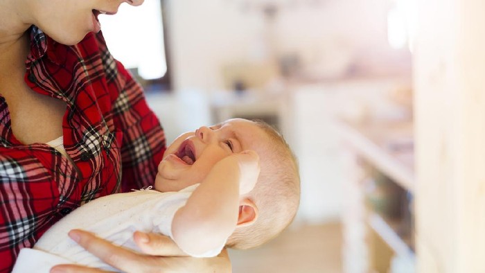 Crying little baby in the arms of her mother in a living room.