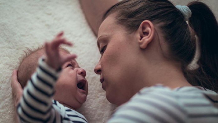 Young single mother holds a baby boy in matching white and blue striped clothes while he is crying. They are lying down on a white blanket.