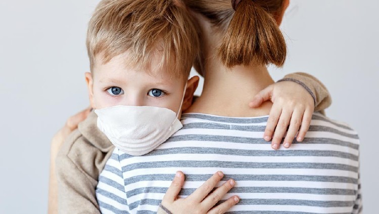 Little boy in medical mask hugging crop mother and looking at camera during coronavirus outbreak against gray background