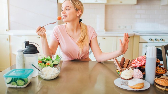 Nice young well-built model eting healthy food in kitchen at table. She says no to junk meal. Alone in kitchen. Daylight. Healthy lifestyle