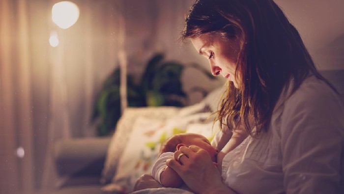 Young beautiful mother, breastfeeding her newborn baby boy at night, dim light. Mom breastfeeding infant