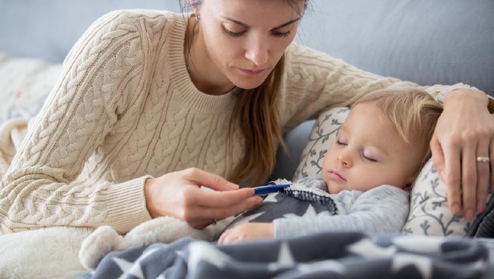 Sick child, toddler boy lying on the couch in living room with a fever, mom cheching his temperature, resting at home
