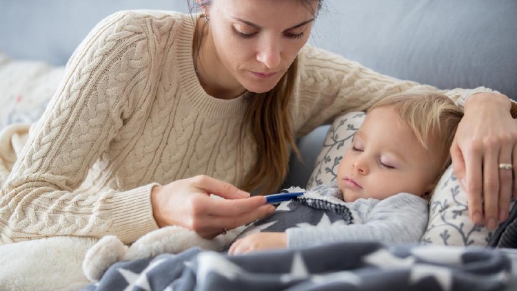 Sick child, toddler boy lying on the couch in living room with a fever, mom cheching his temperature, resting at home