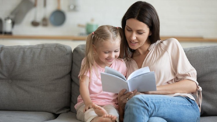 Happy mother and little daughter reading book, sitting on comfortable couch in living room, smiling mum teaching little girl child, family spending weekend at home together, children education