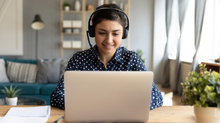 Ilustrasi Kuliah Online Smiling millennial Indian girl in wireless headset sit at desk in living room study online on laptop, happy young ethnic female in headphones watch webinar or course on Internet on computer