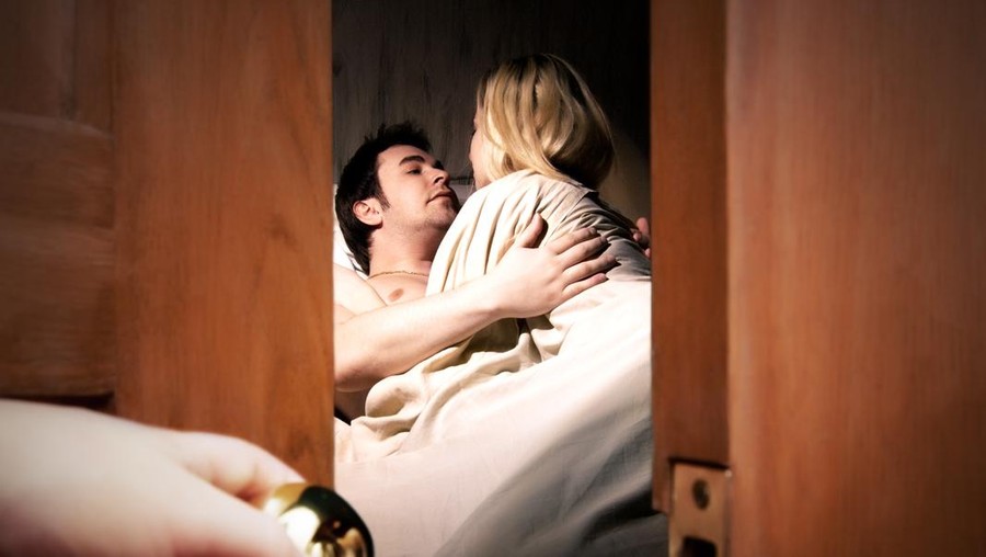 Couple lying in bed while someone is watching in front of the door