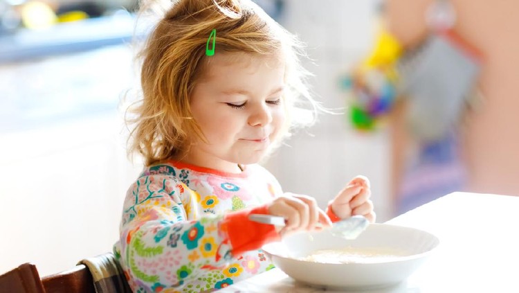 Adorable toddler girl eating healthy porridge from spoon for breakfast. Cute happy baby child in colorful pajamas sitting in kitchen and learning using spoon