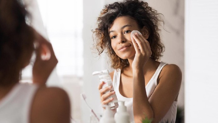 Young afro-american woman cleaning face with cotton pad looking in mirror at bathroom