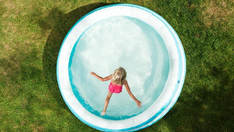 A six year old girl is jumping in an inflatable swimming pool on a hot Summer day. The grass is becoming dry and brown.