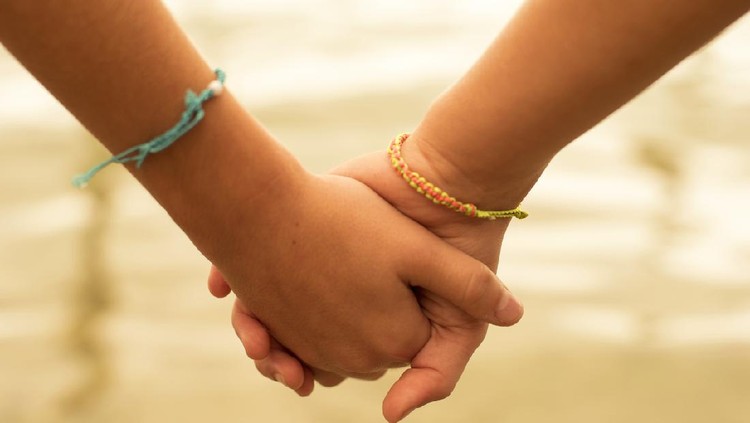 Close up of two children holding hands on the beach sand, summertime friendship concept.