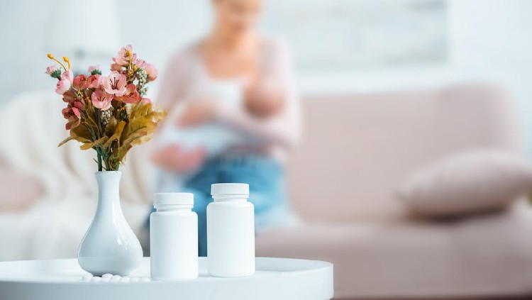 Ibu menyusui containers with pills, flowers in vase and mother breastfeeding baby behind at home