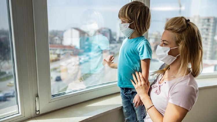 1215892079 Single mother and her small son with face masks looking through window while being in isolation at home. Focus is on boy.