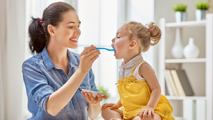 Happy young mother feeding her baby girl with a spoon at home.