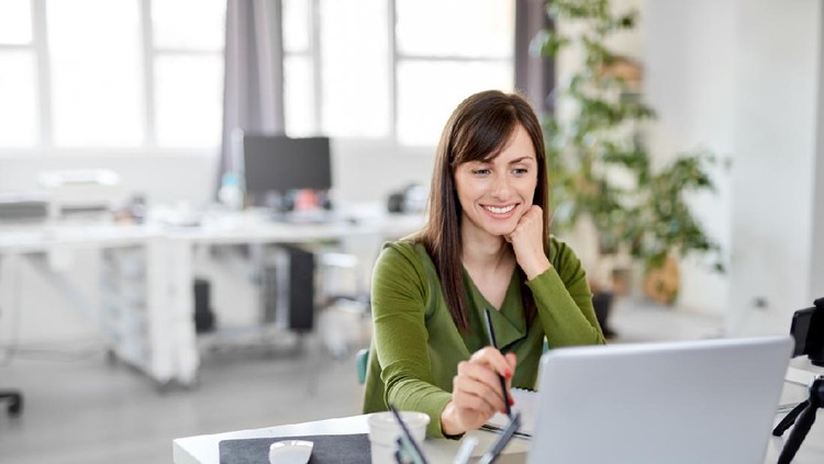 Young Caucasian busy brunette smiling and holding pencil while sitting in office and looking at laptop.
