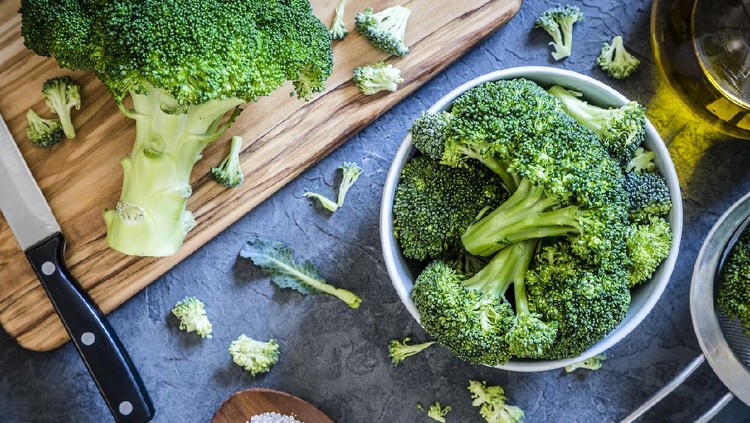 Ilustrasi brokoli Top view of a bowl full of broccoli branches surrounded by a wooden cutting board with a entire broccoli and a kitchen knife on top, an olive oil bottle, a metal colander and a little wooden tray with salt on a grey textured backdrop. Low key DSLR photo taken with Canon EOS 6D Mark II and Canon EF 24-105 mm f/4L