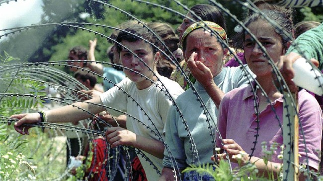 FILE- In this Thursday, July 13, 1995, file picture, refugees from Srebrenica look through the razor-wire at a UN base, outside Tuzla.  Survivors of the genocide in the eastern Bosnian town of Srebrenica, mainly women, will on Saturday July 11, 2020, commemorate the 25th anniversary of the slaughter of their fathers and brothers, husbands and sons.  At least 8,000 mostly Muslim men and boys were chased through woods in and around Srebrenica by Serb troops in what is considered the worst carnage of civilians in Europe since World War II. The slaughter was also the only atrocity of the brutal war that has been confirmed an act of genocide.(AP Photo/Darko Bandic, File)