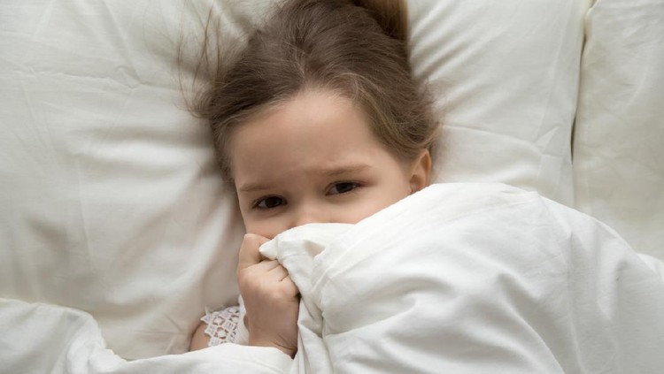 Cute little girl waking up in morning after sleep, covering face with blanket, preschool child lying in comfortable bed with white linens, looking at camera, top view from above close up