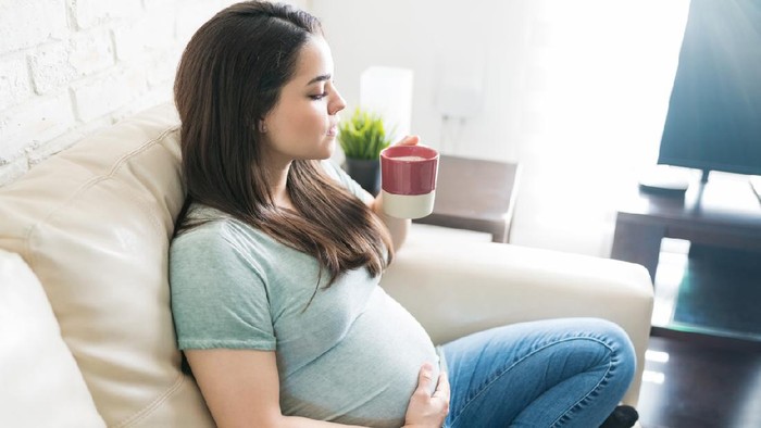 Attractive pregnant female chilling while drinking fresh coffee from mug and relaxing at home