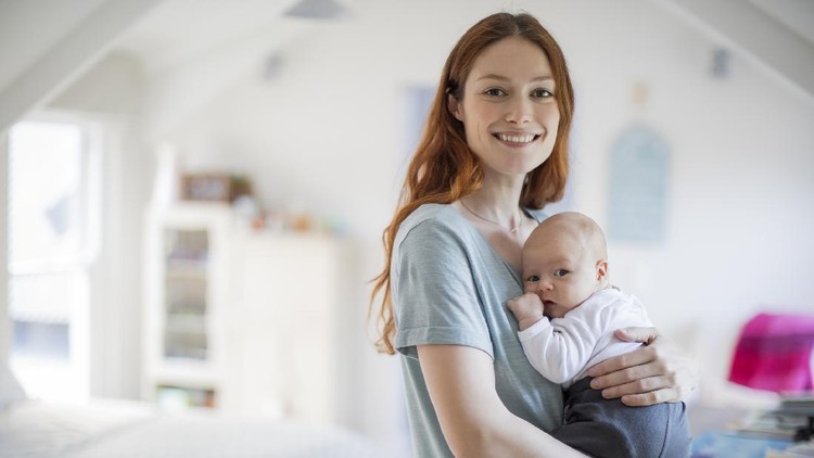 Smiling redhead woman standing with cute son. Portrait of beautiful mother is carrying newborn baby boy in bedroom. They are at home.