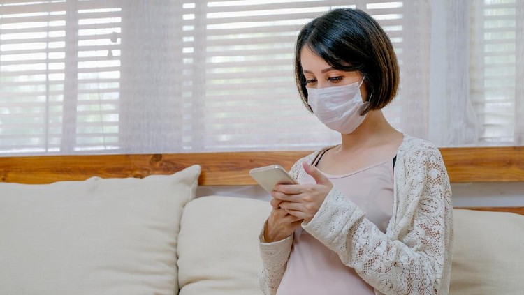 Asian pregnant woman with mask look at mobile phone and sit on sofa in living room of her house with soft light.