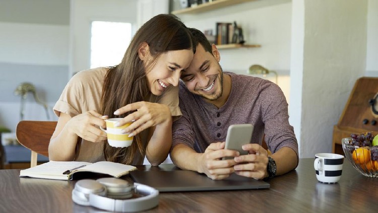 Cheerful boyfriend showing mobile phone to girlfriend in dining room. Young woman is holding coffee cup. They are spending leisure time together at home.