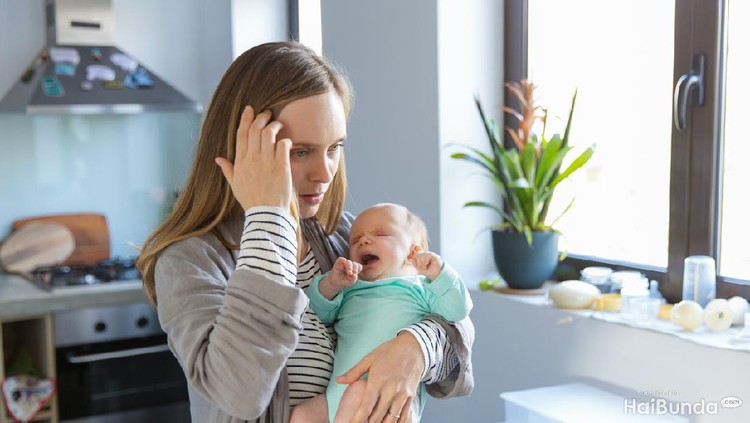 Tired concerned new mother rocking crying baby in kitchen. Portrait of young woman and cute little child in home interior. Motherhood concept