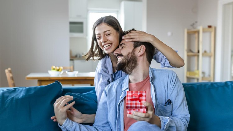 Beautiful Girl Surprise Her Boyfriend With Gift Celebrating Anniversary While He Is Sitting on the Sofa in the Living Room. Romantic Surprise for Husband Boyfriend, Happy Birthday Concept