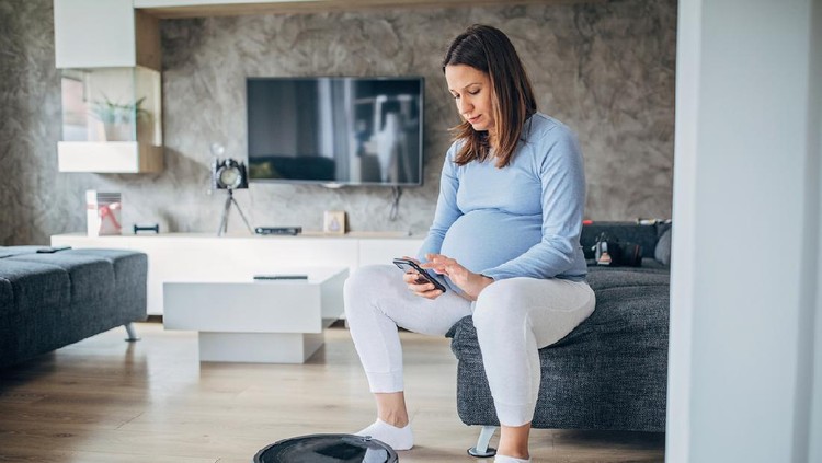 Young smiling pregnant brunette lying in bed with laptop on thighs and using smart phone. Evening time.