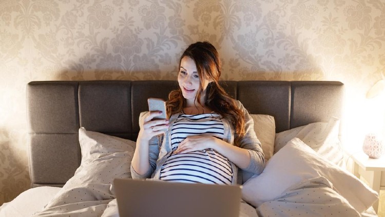 Young smiling pregnant brunette lying in bed with laptop on thighs and using smart phone. Evening time.