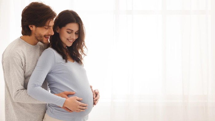 Future parents. Happy expecting couple hugging and smiling while standing near the window at home, free space