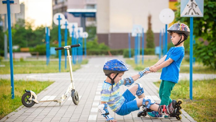 Two boys sibling brothers together in park, helps boy with roller skates to stand up after fall. Friendship and active leisure summer holidays time with family concept.