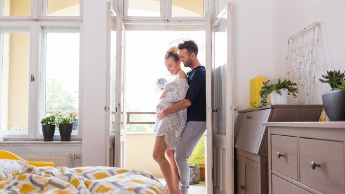 Happy couple in the bedroom in the morning, standing in the open window. Man embracing her pregnant wife.