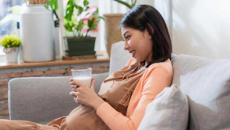 Asian pregnant woman hold a glass of milk for her health and baby with smile and sitting on the sofa, feeling happily and relaxed. Pregnant women concept.