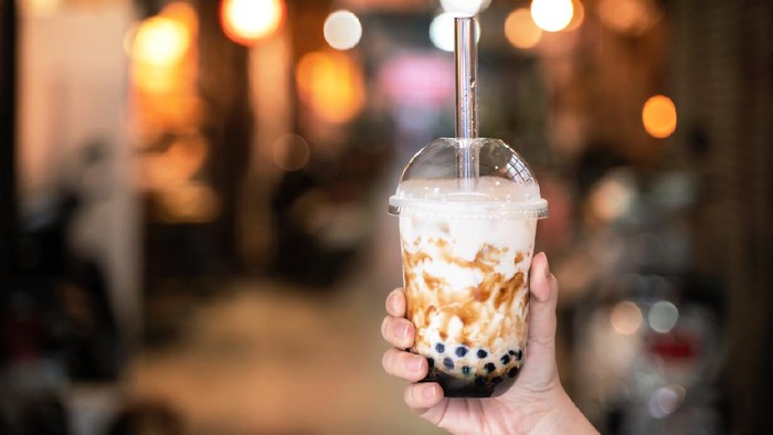 Young girl in denim jacket is drinking brown sugar flavored tapioca pearl bubble milk tea with glass straw in night market of Taiwan, close up, bokeh