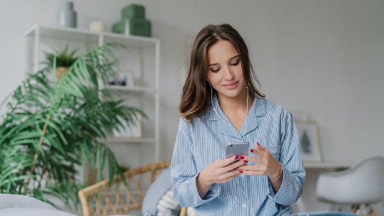 Photo of relaxed young European lady in pyjamas enjoys audio playlist, listens music romantic songs in earphones, uses modern cell phone, sits on comfortable bed against domestic atmosphere.