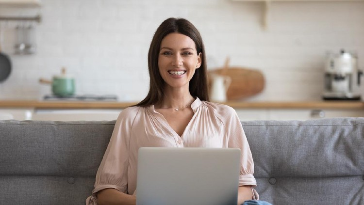 Portrait of smiling businesswoman sitting alone on comfortable sofa at home with computer on knees. Happy female freelancer working remotely in own house, posing for photo, looking at camera.