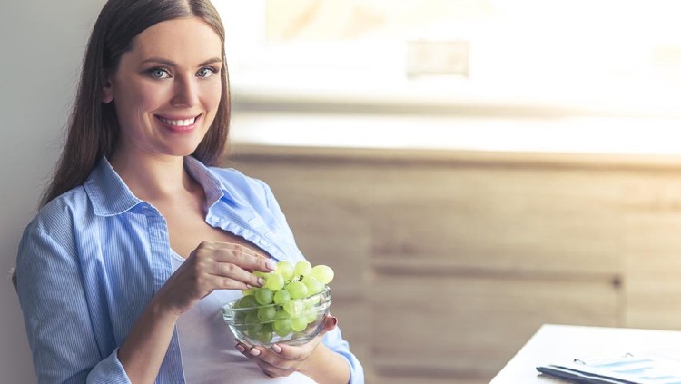 Beautiful pregnant business woman is eating grape, looking at camera and smiling while sitting at her working place at home
