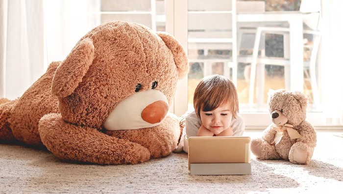 Cute little child watching cartoons on digital tablet device lying on floor with two soft teddy bear toys at home. Modern childhood.
