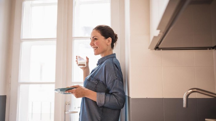 Young woman eating a croissant, drinking fresh milk for breakfast in the kitchen. She is wearing only a blue shirt, standing by the big, bright window