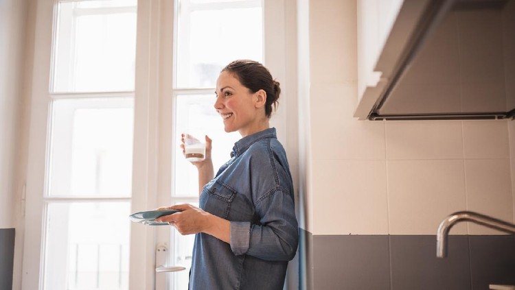 Ilustrasi wanita minum susu Young woman eating a croissant, drinking fresh milk for breakfast in the kitchen. She is wearing only a blue shirt, standing by the big, bright window