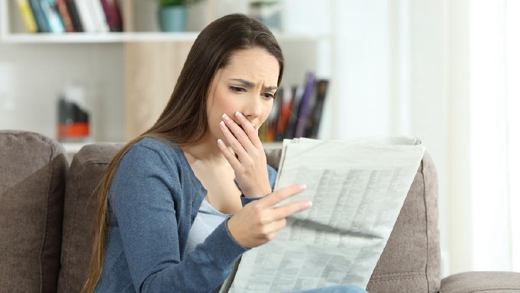 Ilustrasi membaca Worried woman reading bad news in a newspaper sitting on a couch in the living room at home